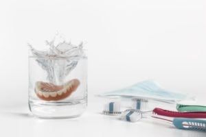 Dentures in cup of solution next to multiple tooth brushes